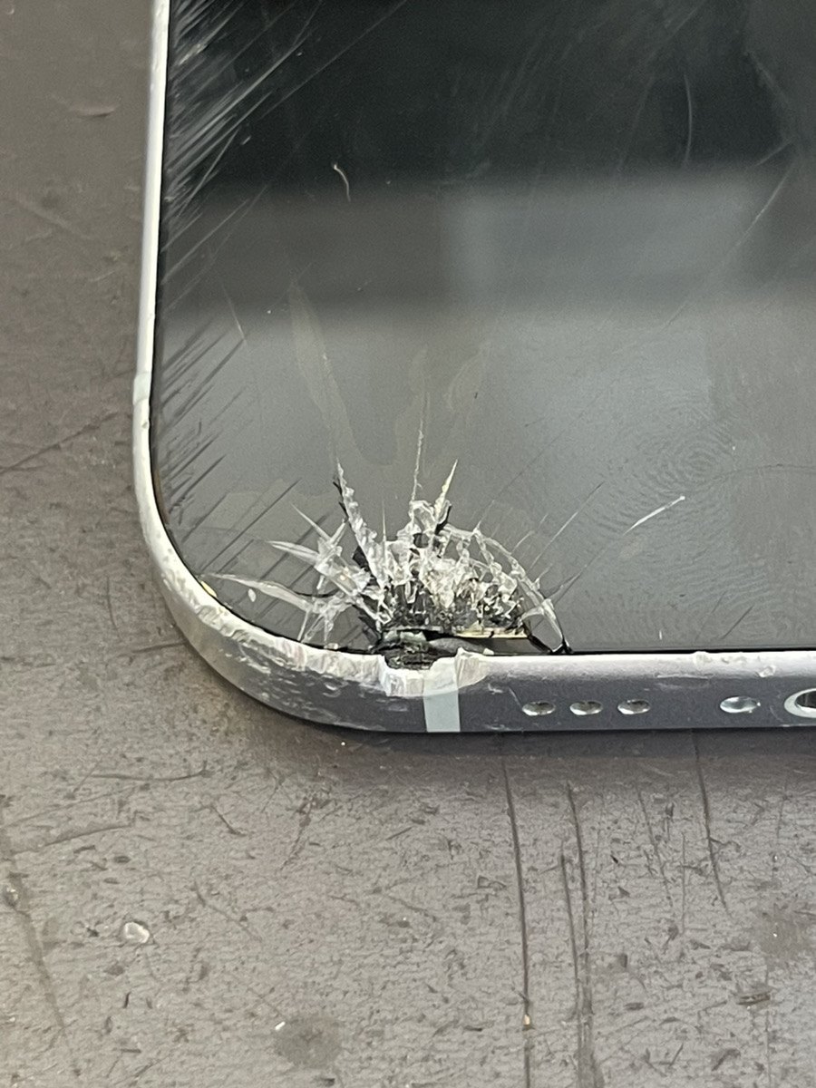 iPhone 15の左下の角。ガラスが放射状に割れ、アルミのフレームが内側に潰れて画面に食い込んでいる