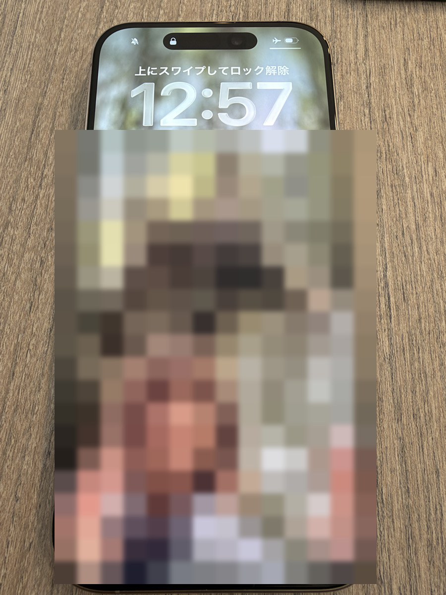 修理後に起動したiPhone 15 Proのロック画面。時計が表示され、機内モードのアイコンが点いている。壁紙はモザイク処理